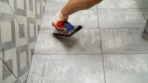 Close up of worker in protective glove applying grout to grey ceramic floor tiles with float tool during bathroom renovation