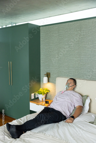 Senior Chinese man reclining on bed wearing oxygen mask and tubing near nightstand with smartphone
