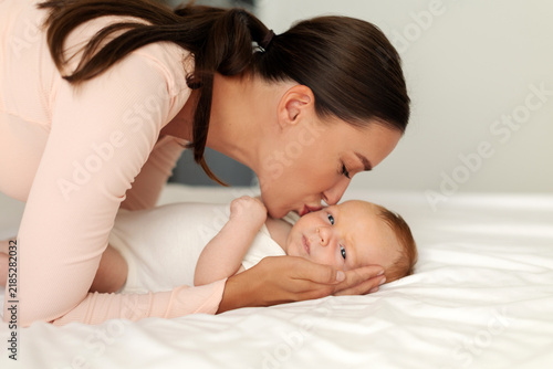Fotografie Mother lovingly kissing newborn baby lying on bed, tender side view portrait