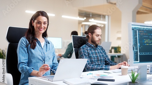 Camera approaching young Caucasian beautiful happy woman in glasses typing on laptop looking at camera and smiling. Professional people work in modern office. Business. Close up. Worker concept