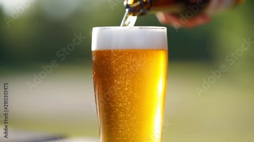 Slow motion close up static of beer being poured into pint glass on outdoor table in garden during daytime summer warm light