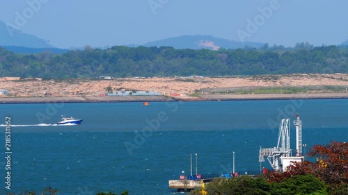 Fast boat cruising across blue coastal waters in Singapore with industrial pier dock in foreground and sandy coastline in background on clear sunny day