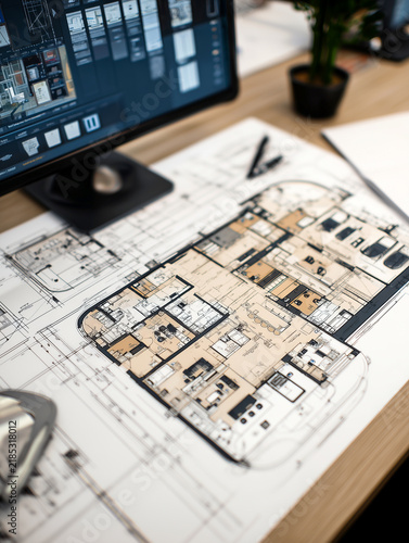 Detailed architectural blueprints and house floor plans lie spread out on a wooden office desk next to a computer monitor showing design software for building construction