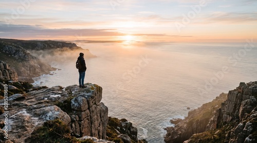 Lone hiker stands on cliff edge overlooking misty ocean at sunrise with sunbeams breaking through clouds