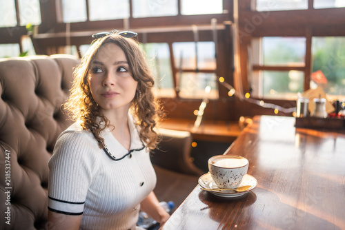 Young curly woman sitting with coffee in sunny cafe looking away