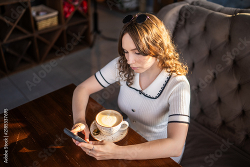Young woman with curly hair using smartphone with coffee in cafe