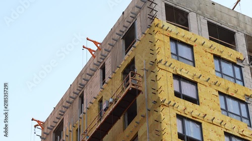 Construction workers installing exterior insulation and facade materials on a modern building using a suspended platform, showing high rise construction, energy efficiency and professional facade work