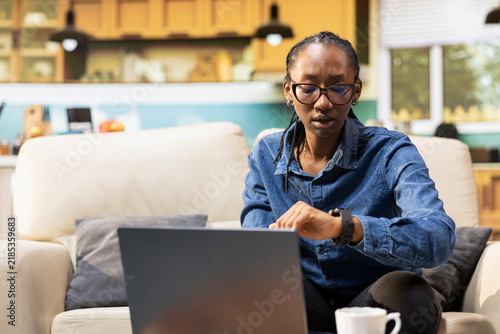 Quadro su tela African American girl checks the time on her wristwatch for time management, punctual and anxious about a teleworking schedule; young digital nomad enjoys domestic life and punctuality at home
