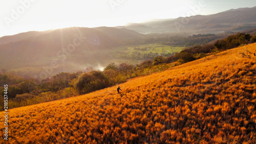 Canvas Print A lone hiker walking across a golden grass-covered hillside overlooking a green valley and distant mountain ranges under warm natural sunlight