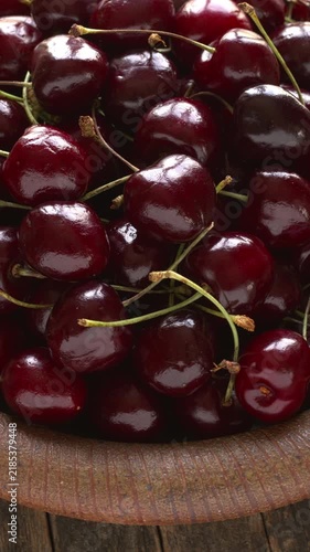 Sweet cherries with stems rotating in close-up on a handmade dish. Fresh organic red fruits heaped on a ceramic plate on a dark, rustic wood background. Vertical video.