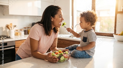 Happy toddler boy feeding mother fresh salad in sunny kitchen