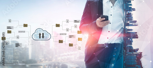 Картината върху платно A businessman stands confidently, engrossed in his smartphone, with a city skyline and cloud technology overlay, symbolizing modern finance and digital connectivity