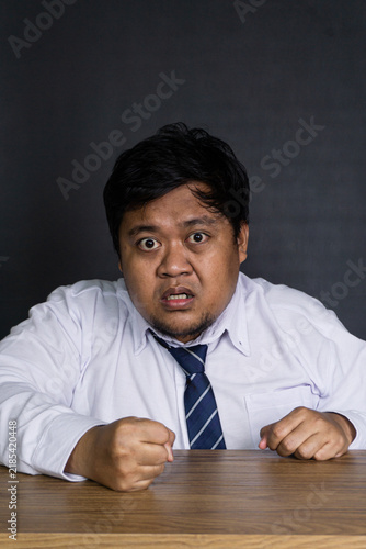 Foto An infuriated young Asian male professional leans aggressively forward at a smooth wooden desk against a plain dark gray wall, with both fists clenched tightly on the surface