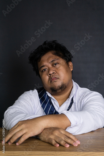 Foto A highly unmotivated young Asian male corporate employee leans forward at a smooth wooden desk against a solid dark gray wall, resting both arms comfortably on the surface