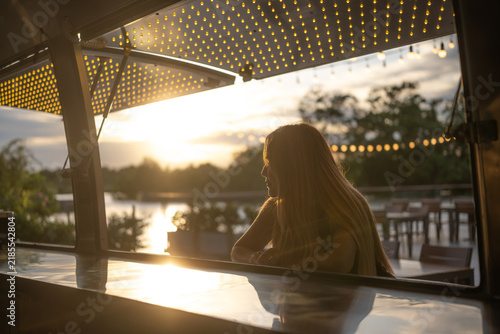 A woman sat at a food counter by the river in the evening.