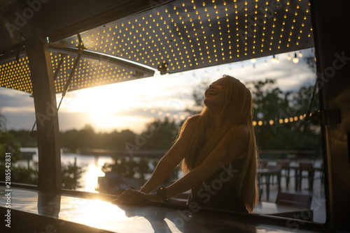 A woman sat laughing happily at a food counter by the river in the evening.