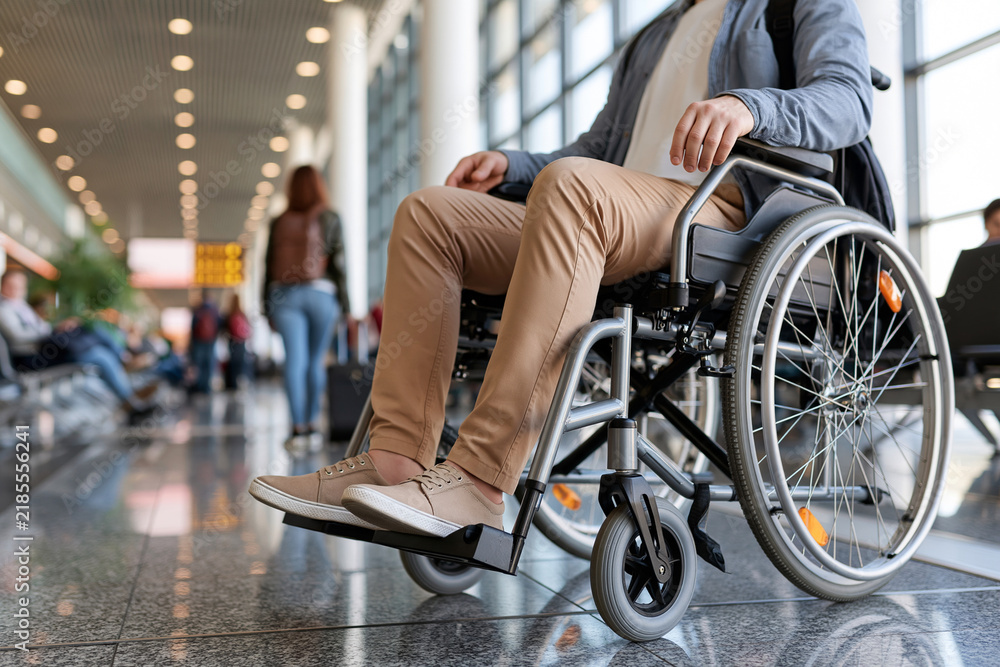 Obraz premium Person in wheelchair moves through airport terminal. Shoes, trousers, and jacket match neutral tones. Background shows passengers waiting at distant gates. Glass ceiling reflects ambient lights above