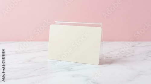 Blank white business card in clear acrylic stand on marble surface, pastel pink background, minimalist corporate mockup, office branding concept with copy space, professional stationery template