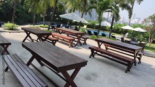 Sunlit patio holds rustic wooden tables and benches. Tropical palms sway gently in the breeze. White umbrellas shade the open-air seating area. Calm pool water reflects the bright sky