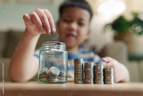 Quadro su tela Asian boy putting a coin into a savings jar beside growing stacks of coins, representing financial education, money saving, budgeting, future planning, wealth growth and responsibility