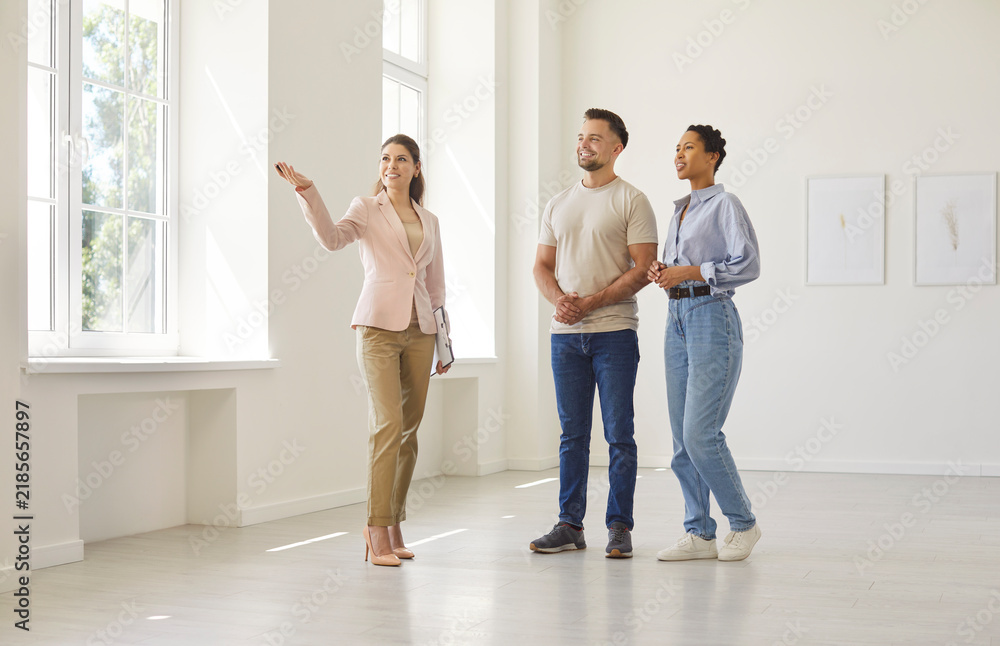 Fototapeta premium Professional real estate agent guides a young couple on a property tour through a bright empty apartment, briefly explaining layout and features. Realtor shows new house to happy clients.