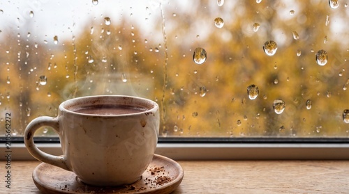 Hot cup of coffee on a wooden windowsill with rain drop on the glass and blurred autumn background