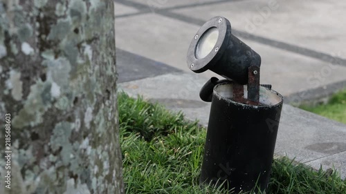 Black spotlight stands near tree trunk and grass. Light fixture mounted on metal base beside paved path. Grass edges and stone slab surround the fixture
