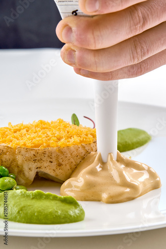 Chef applying sauce with a piping bag onto a gourmet fish dish with green puree in a white plate. High-end dining prep.