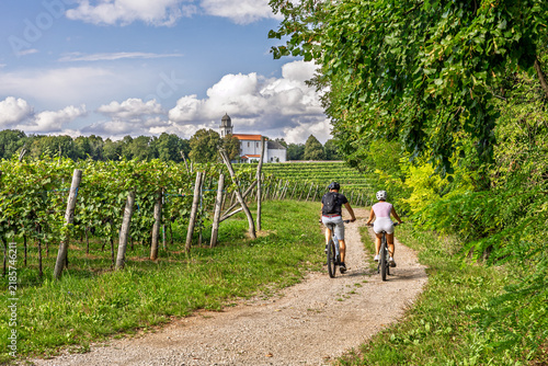 Couple Cycling Through Vineyard Toward Rural Church in Karst
