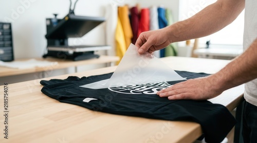 Man's hands peeling back a transfer sheet from a custom t-shirt after heat pressing in a workshop.