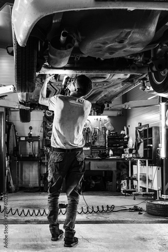 Black and white photo of a mechanic inspecting the underside of a raised car in a busy auto repair garage with tools, equipment, and workshop details visible