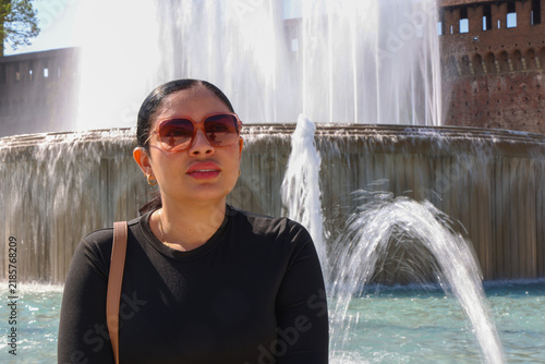 Cuadro en lienzo A close-up outdoor portrait of a dark-haired woman wearing tinted sunglasses and a black top, posing gracefully in front of a large, splashing municipal fountain