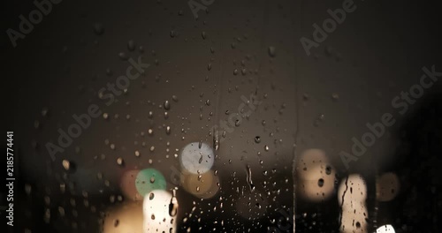 Water drops and rain streams on window pane illuminated by frequent lightning flashes during a night thunderstorm with out-of-focus city lights in bokeh background in real time video.