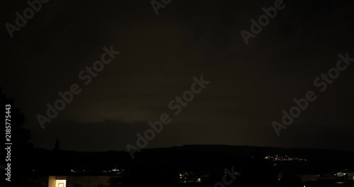 Time lapse of heavy night storm clouds illuminated by bright lightning flashes above a hillside village and residential area.