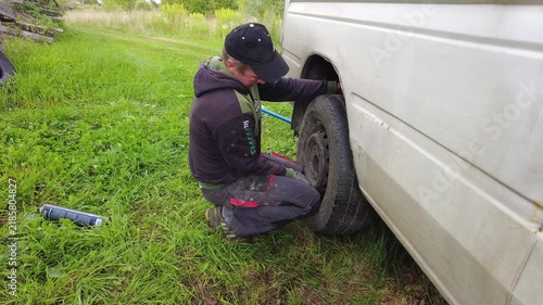 A person is kneeling down and removing a tire from a white van. The setting is an open grassy area with trees in the background. Tools are placed nearby as the person focuses on fixing the tire. 