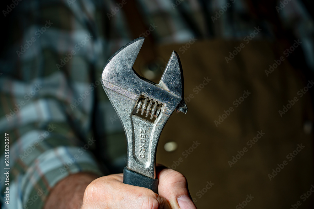 Obraz premium Closeup of hand holding heavy adjustable wrench. Mechanic or carpenter hand gripping vintage steel spanner repair worker with industrial hand tool wearing protective apron background