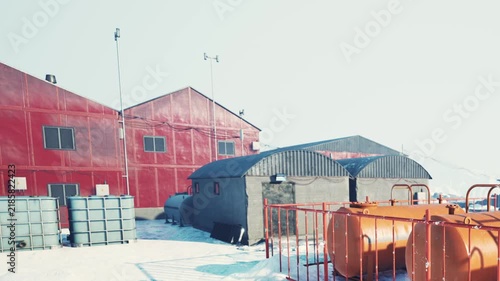 People are working at a research station surrounded by snow. There are red and gray buildings, large tanks, and a clear sky above them, showing the cold environment of the setting.
