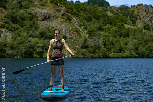 Woman smiles standing on blue stand up paddleboard on lake with trees on shore in background holding paddle and wearing two piece swimming costume with shorts; lady on SUP board balancing having fun