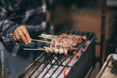 Принти на полотні Close-up of a street food vendor grilling skewers of chicken meat and entrails over glowing hot charcoal on a smoky grill at an outdoor market