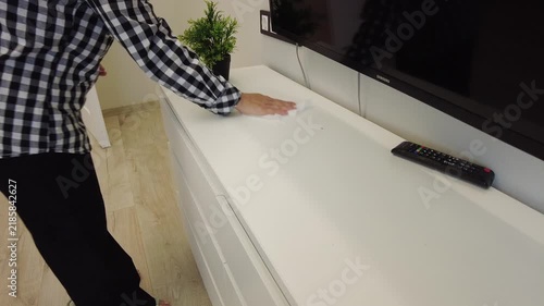 A person is wiping a white shelf in a living room. The shelf is near a television and has a small plant on it. The individual is wearing a black and white checkered shirt. Light enters the room from 