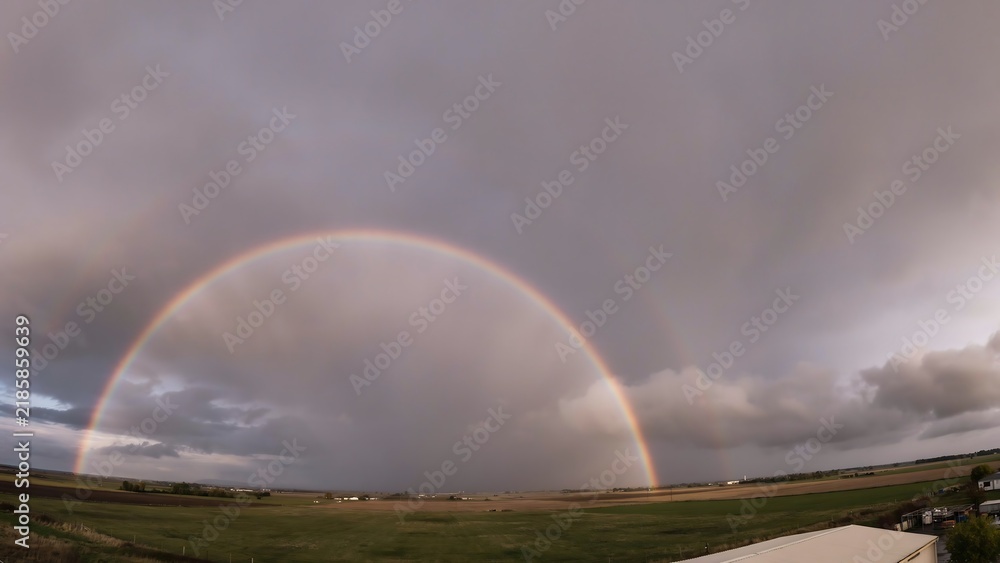 Obraz premium Rainbow arches over a wide green field under a cloudy sky.