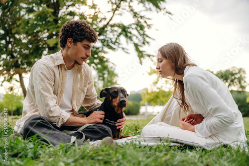 Fotografie Young couple sitting on grass in a park, interacting with a black dog, surrounde