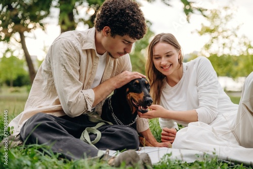 Фотография Young couple sitting on grass in a park, interacting with a black dog, surrounde