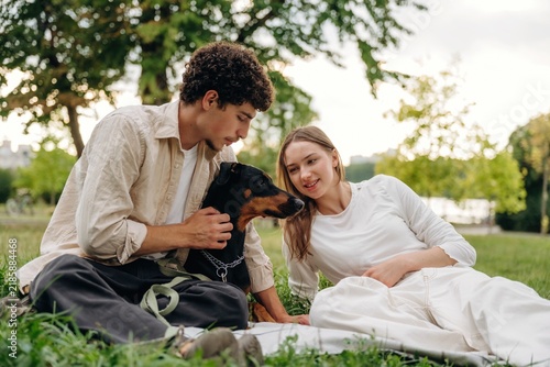 Фотография Young couple sitting on grass in a park, interacting with a black and tan dog, s
