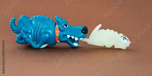 Konstfotografi A playful blue cartoon dog-dinosaur toy chasing or biting a small glow-in-the-dark creature, possibly a rodent or a monster, on a simple brown background