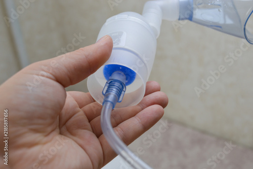 Woman hand supporting the bottom of a nebulizer medicine chamber showing the blue air tube connection close-up. Respiratory equipment assembly, air hose setup, and home healthcare concept