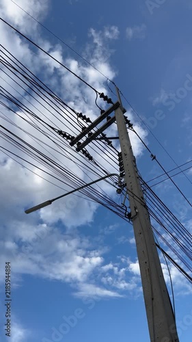 Static vertical shot of a concrete utility pole and overhead power lines against a blue sky with scattered clouds.