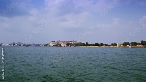 A broad lake-level view looks toward Udaipur's City Palace complex and low waterfront walls beneath a lightly clouded sky.