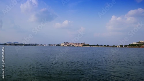 Wide view from the lakefront toward Udaipur's distant City Palace, with blue water and scattered clouds filling the scene.
