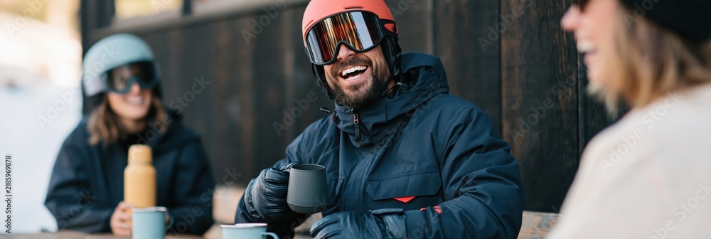 Obraz premium Male skier laughing at outdoor lodge table with friends holding mugs for après ski winter background and lifestyle